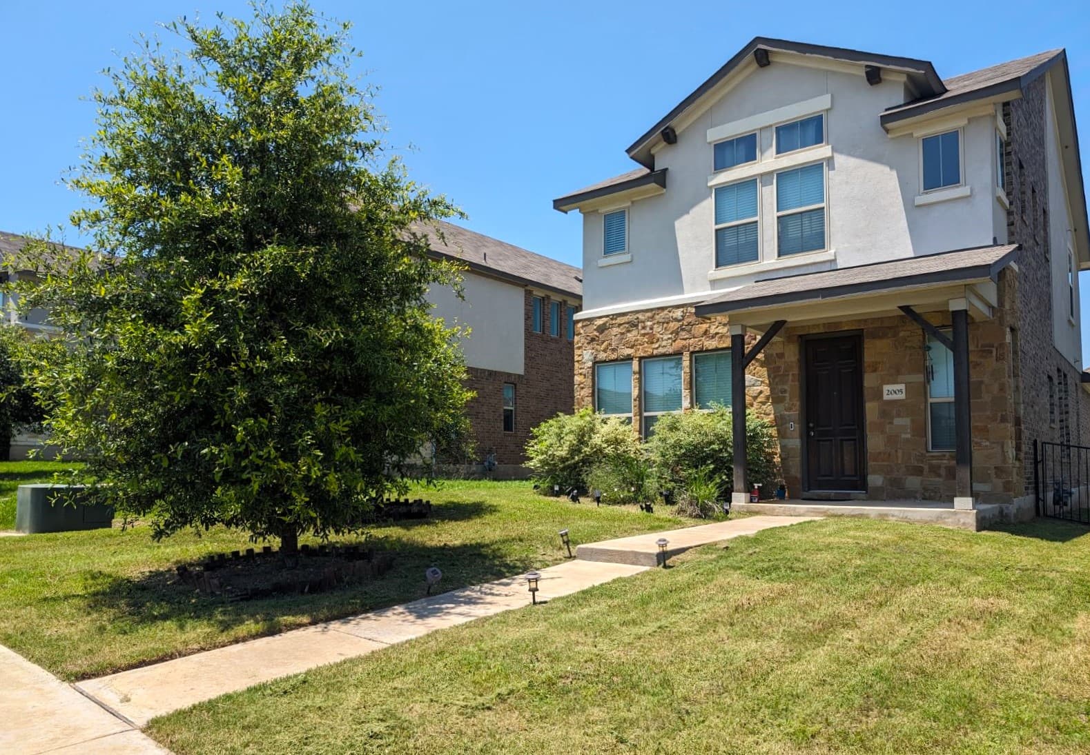 Modern two-story house with stone facade, front porch, green lawn, and a large leafy tree.